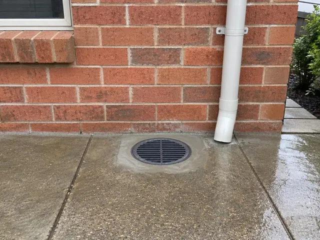 Clean outdoor drain grate set in wet concrete paving beside a brick wall in Charlestown